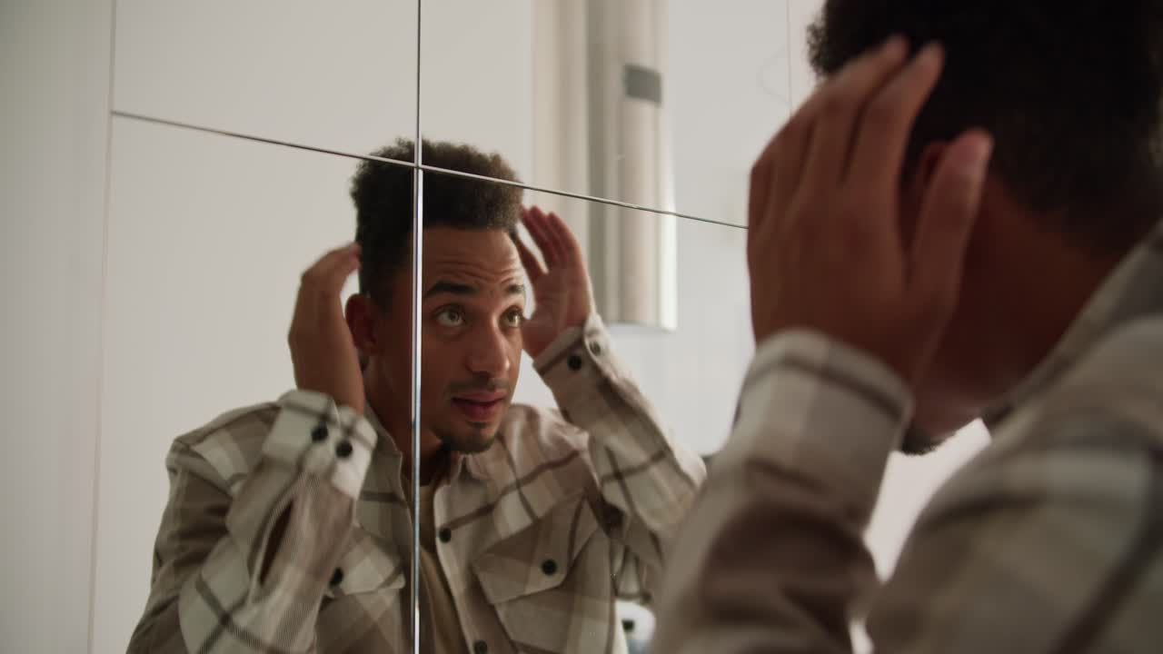A young Black-skinned brunette man with stubble in a cream-colored checkered shirt preens near a mirror in the hallway in a modern apartment with light walls and good lighting