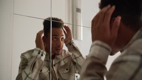 a young black-skinned brunette man with stubble in a cream-colored checkered shirt preens near a mirror in the hallway in a modern apartment with light walls and good lighting