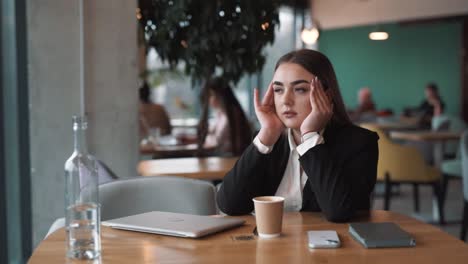 tired,-beautiful-woman-sits-in-a-stylish-cafe-in-business-attire,-closing-her-laptop-and-finishing-work-with-a-headache,-massaging-her-temples-to-relieve-migraine-pain