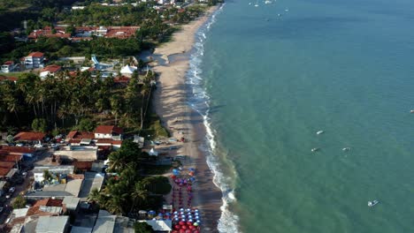 Dolly-out-bird's-eye-drone-shot-of-Penha-beach-coastline-near-the-capital-city-of-Joao-Pessoa-in-Paraiba,-Brazil-with-colorful-umbrellas,-waves-crashing-into-the-sand-and-small-fishing-boats-docked