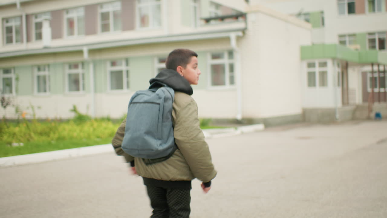 흐린 하늘 아래 학교 건물을 향해 옆을 흘끗 보며 달려가는 배낭을 멘 남자아이가 도시 주거 건축물과 녹지로 둘러싸여 결연함과 활력을 보이는 모습