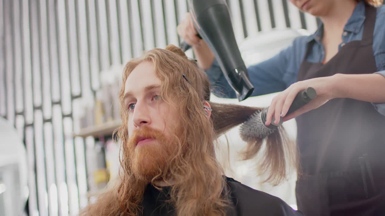 Caucasian female hairdresser styling male client's long hair with hairdryer and brush, slow motion