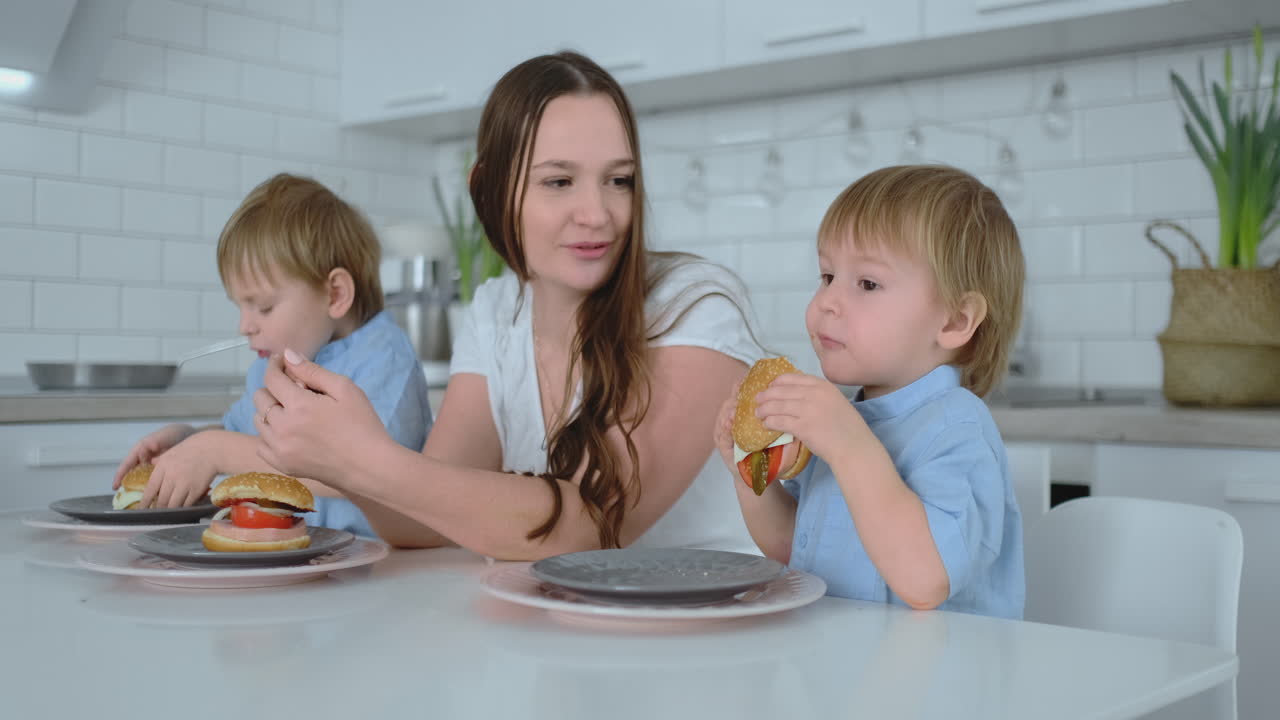 bebé sentado en la cocina con su madre y su hermano comiendo una hamburguesa y sonriendo. comida saludable, hamburguesas caseras