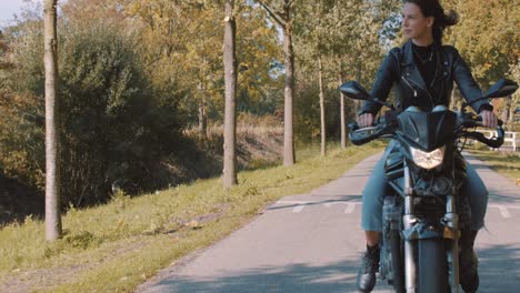 Pretty-smiling-European-young-woman-driving-a-motorbike-wearing-leather-jacket-in-forest-with-vibrant,-colorful-golden-autumn-leaves-on-sunny-day