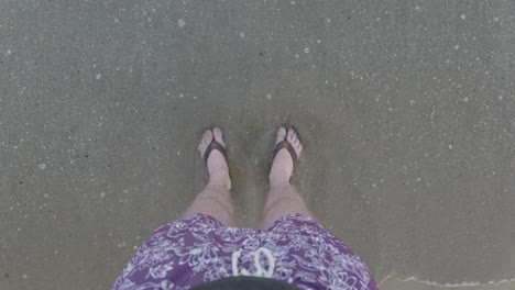 men feet in sandals stand on the beach - waves come easily to the beach and cover the feet