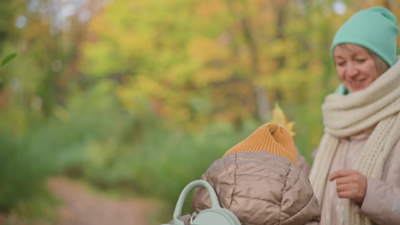 tender moment as mother leans in to kiss daughter on forehead while both wear cozy winter hats and jackets, surrounded by soft-focus fall foliage in peaceful forest setting