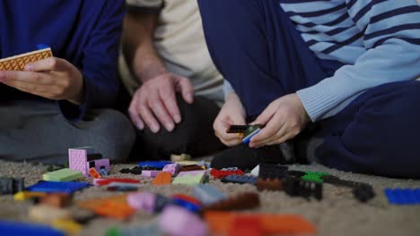 Family are folding the multi-colored blocks of the constructor in different buildings and models of transport on the carpet in the room. Close-up