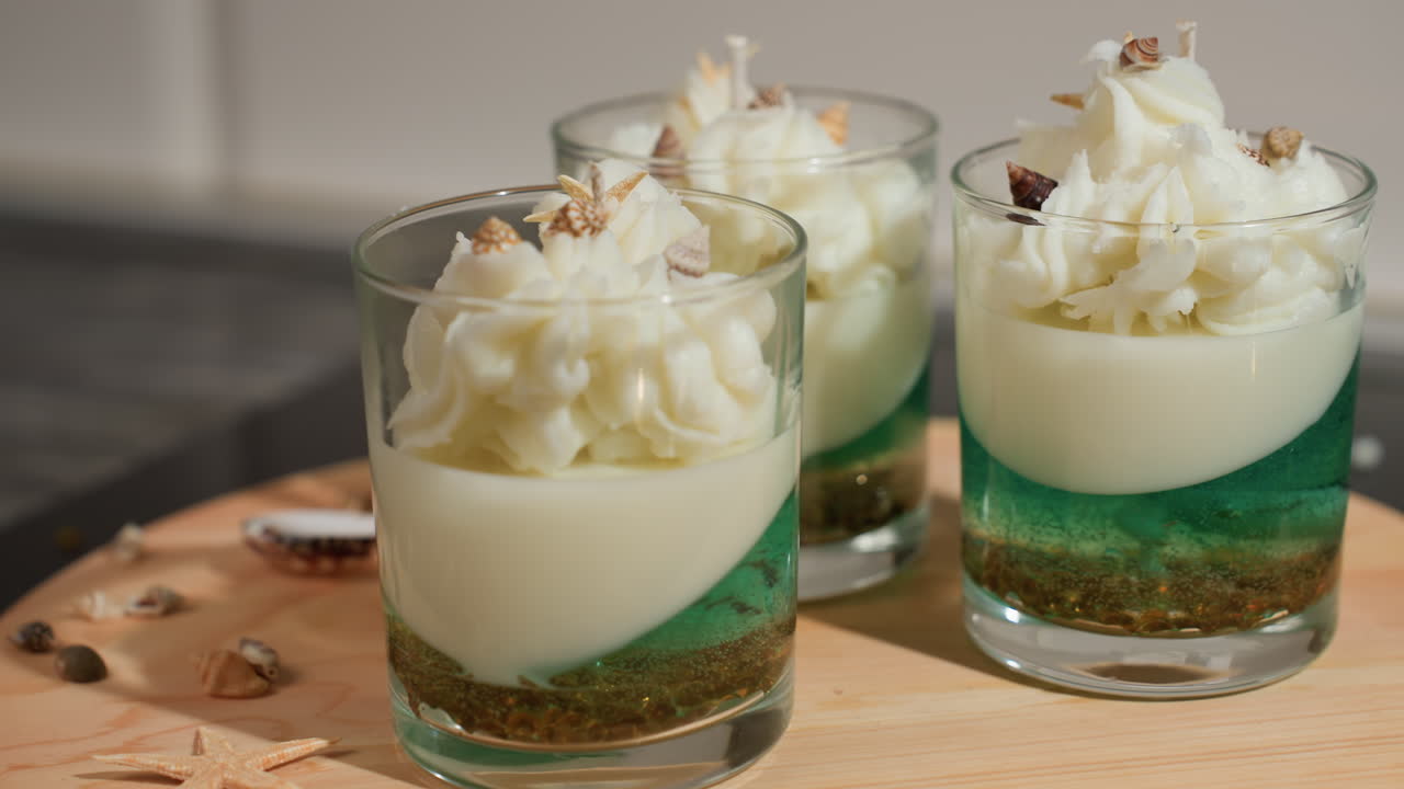 Close up of well decorated parfait candle in transparent glass, featuring green and white wax layers topped with whipped wax swirls and miniature shell accents, placed on round wooden tray