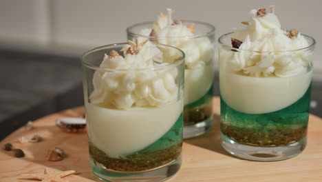 close up of well decorated parfait candle in transparent glass, featuring green and white wax layers topped with whipped wax swirls and miniature shell accents, placed on round wooden tray