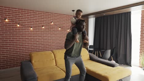 young dad does squats in cozy living room, his toddler son sits on shoulders