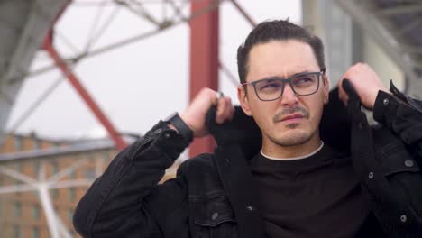 A-serious-young-man-with-glasses-adjusting-his-collar-on-a-windy,-overcast-day