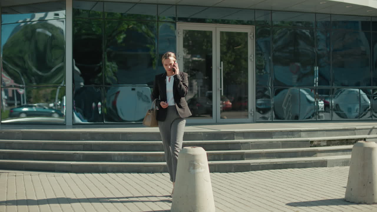 Banker on phone call stepping down staircase outside modern glass office building, holding handbag with sleek reflections of cars and surroundings on facade