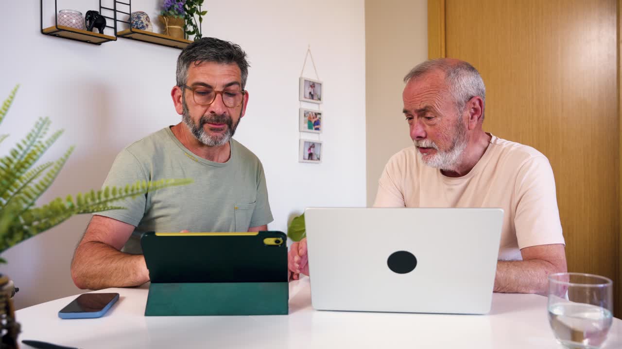 Two Men Discussing Content on Laptop and Tablet at Home