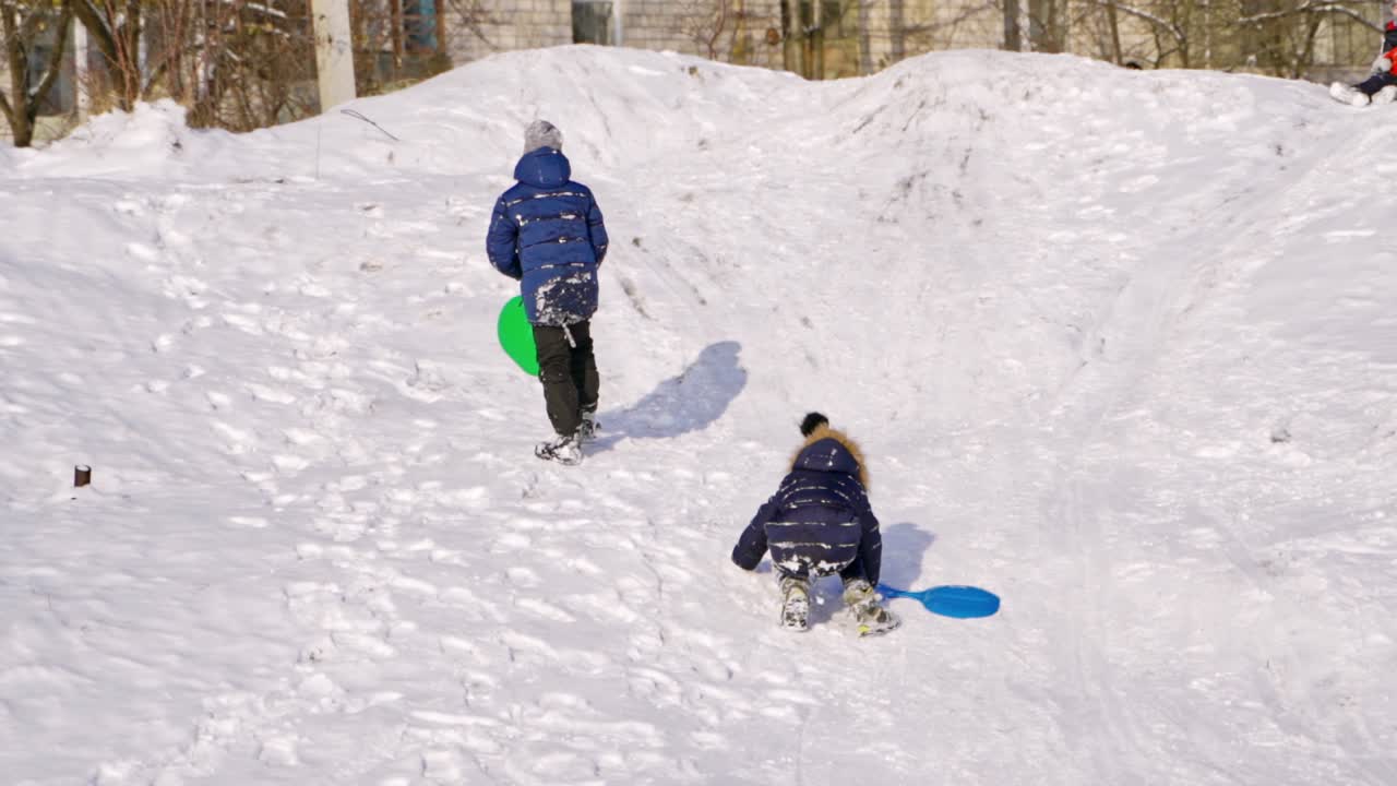 Boys with plastic sleds on a slide in winter. Back view of children on snow background. Slow motion of boys rising up the snowy hill outdoors.