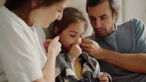 Two-parents,-a-brunette-man-in-a-gray-T-shirt-and-his-wife,-a-brunette-girl-in-a-white-T-shirt,-are-caring-for-and-stroking-their-sick-daughter-who-is-sitting-in-a-blanket-on-the-sofa