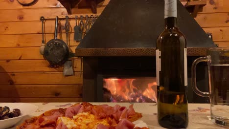 extender la mano para recoger una copa de vino de la mesa junto a una pizza rústica con chimenea de fondo en una cabaña de madera