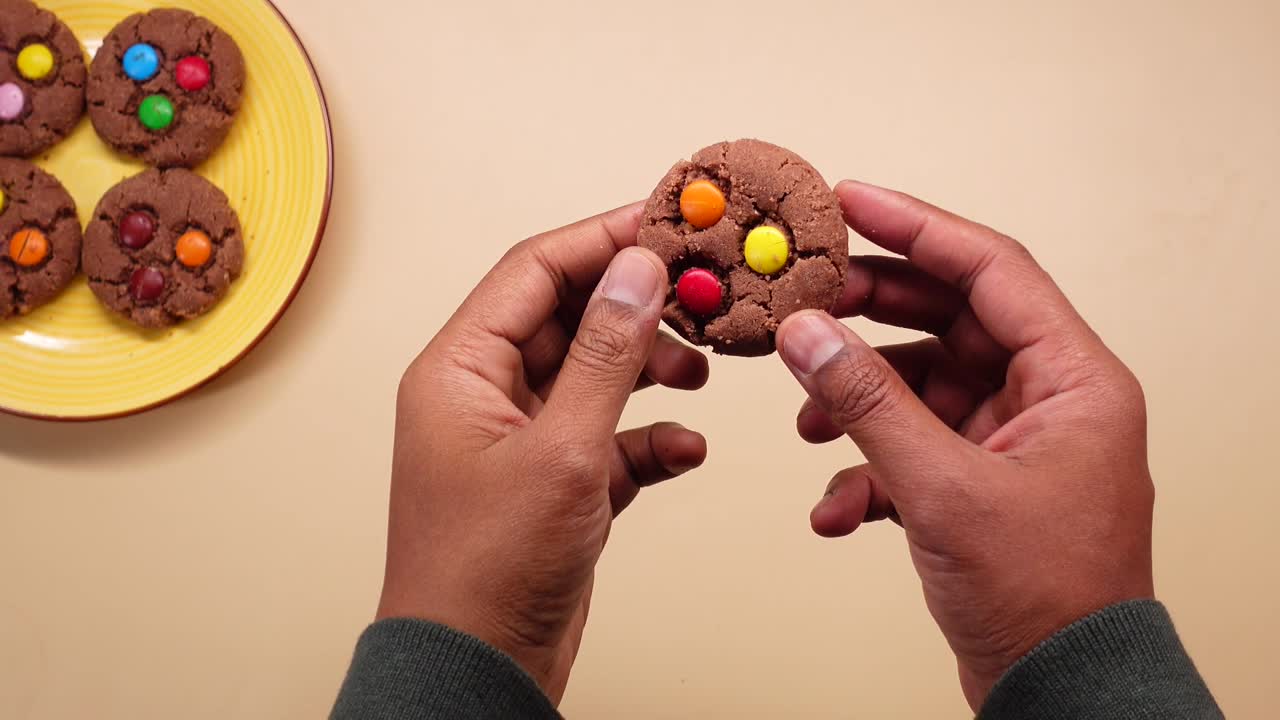 Cận cảnh từ trên cao: Bàn tay đang bẻ một chiếc bánh quy sô cô la với kẹo đầy màu sắc