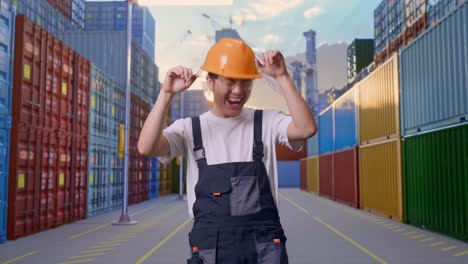 un trabajador asiático con gafas de protección y casco de seguridad bailando mientras está de pie en un almacén de contenedores