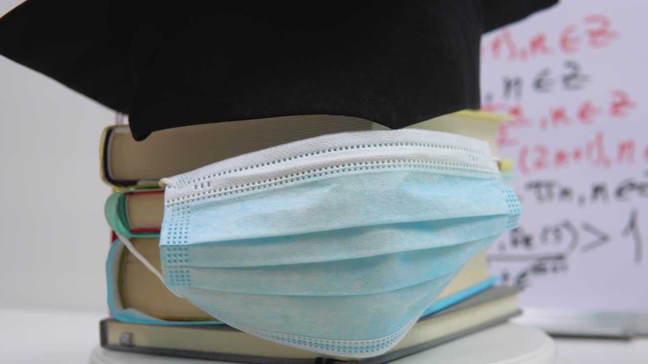 주 (主) 의 검은 사각형 모자는 교과서 위에 놓여 있으며, 기둥으로 쌓여 있으며, 보호 의료 마스크로 장식되어 있습니다. 대학 졸업생의 속성은 그 축을 중심으로 스탠드 위에 회전합니다.