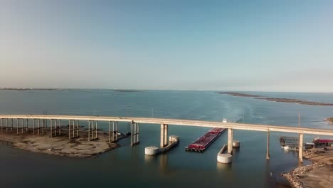 Aerial-view-of-large-red-barge-traveling-under-the-JFK-Causeway-at-southern-end-of-Corpus-Christi-Bay-in-the-Gulf-Intercoastal-Waterway-channel-in-Texasi-Texas