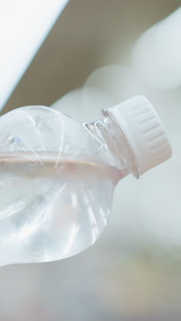 dama sostiene agua embotellada fría con condensación visible en la superficie, en un centro comercial bien iluminado, mientras que el fondo presenta a otras personas caminando, borrosa, la botella es clara y fría