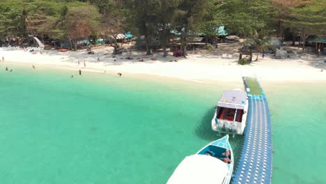 Tracking-of-Koh-Hey-shoreline-with-tourists-enjoying-the-emerald-waters-and-moored-boats-on-floating-piers,-in-Thailand---Aerial-low-angle-fly-over-shot