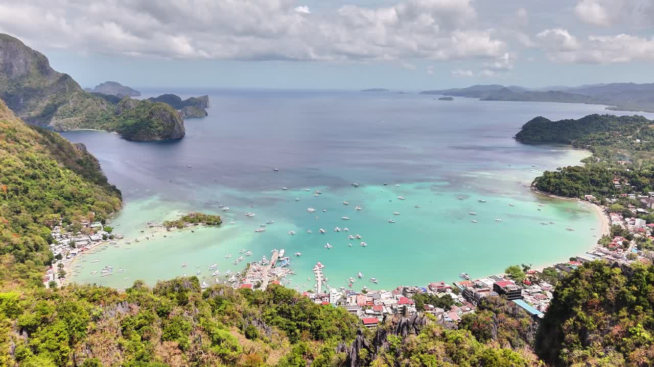 Turquoise waters meet limestone cliffs in this coastal bay scene filled with boats and surrounded by lush greenery and scattered buildings under a partly cloudy sky
