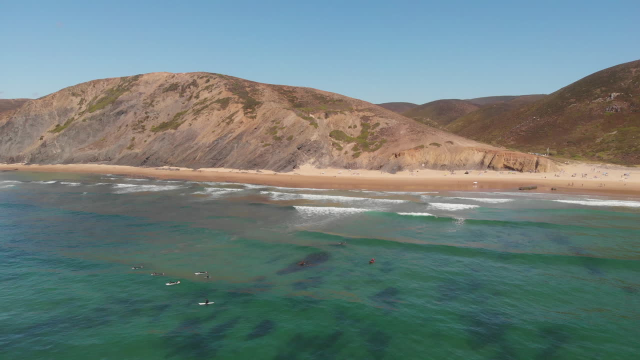 Aerial: Wide high angle view of a group of surfers resting on their surfboards while high waves reach the shore and hilly land is revealed in the background.