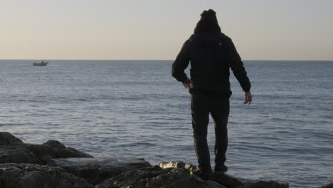 man-walks-onto-stone-terrain-to-gaze-at-fishing-boat-sailing-on-sea,-during-morning-sun