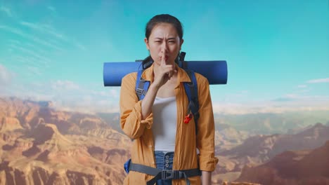 asian female hiker with mountaineering backpack looking at camera and making shh gesture while traveling at the top of mountain