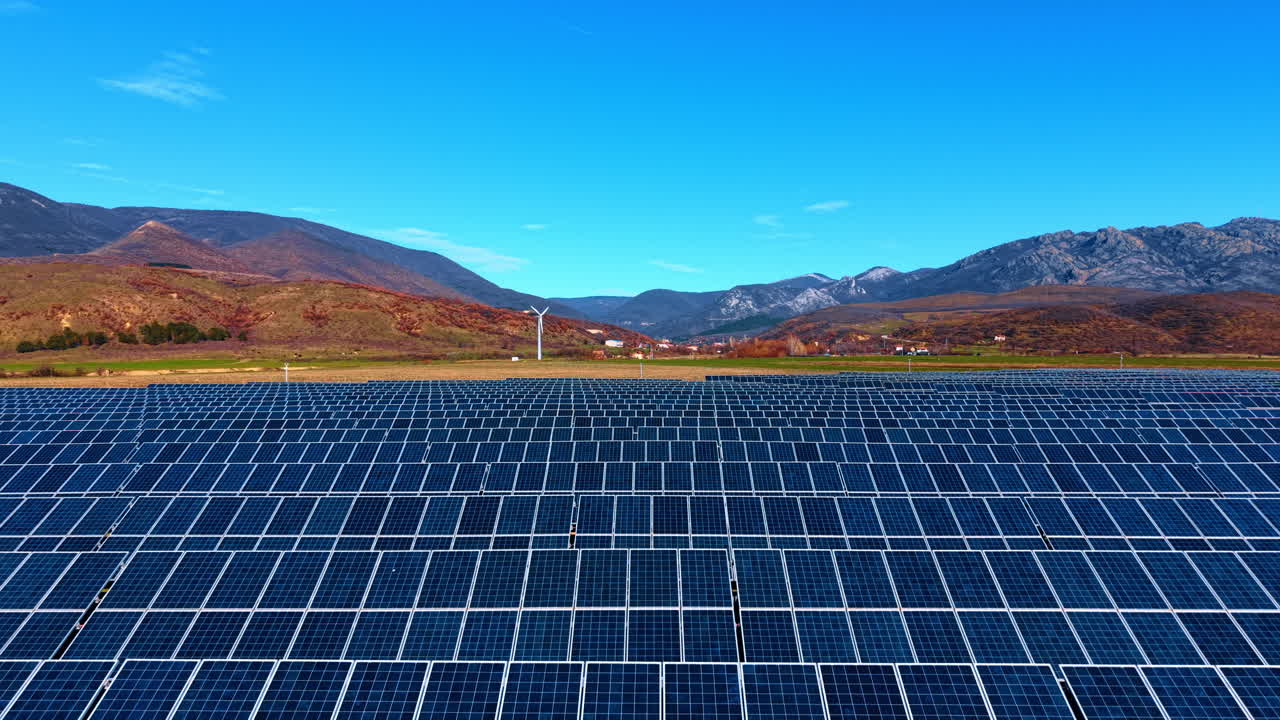 風力タービンと山々を背景にした大規模なソーラーパネル発電所。山々と風力発電機のある澄み渡る青空の下、広がる太陽光発電所の列のワイドな風景