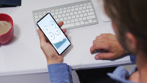 hombre caucásico sentado en un escritorio usando un teléfono inteligente con estadísticas en la pantalla