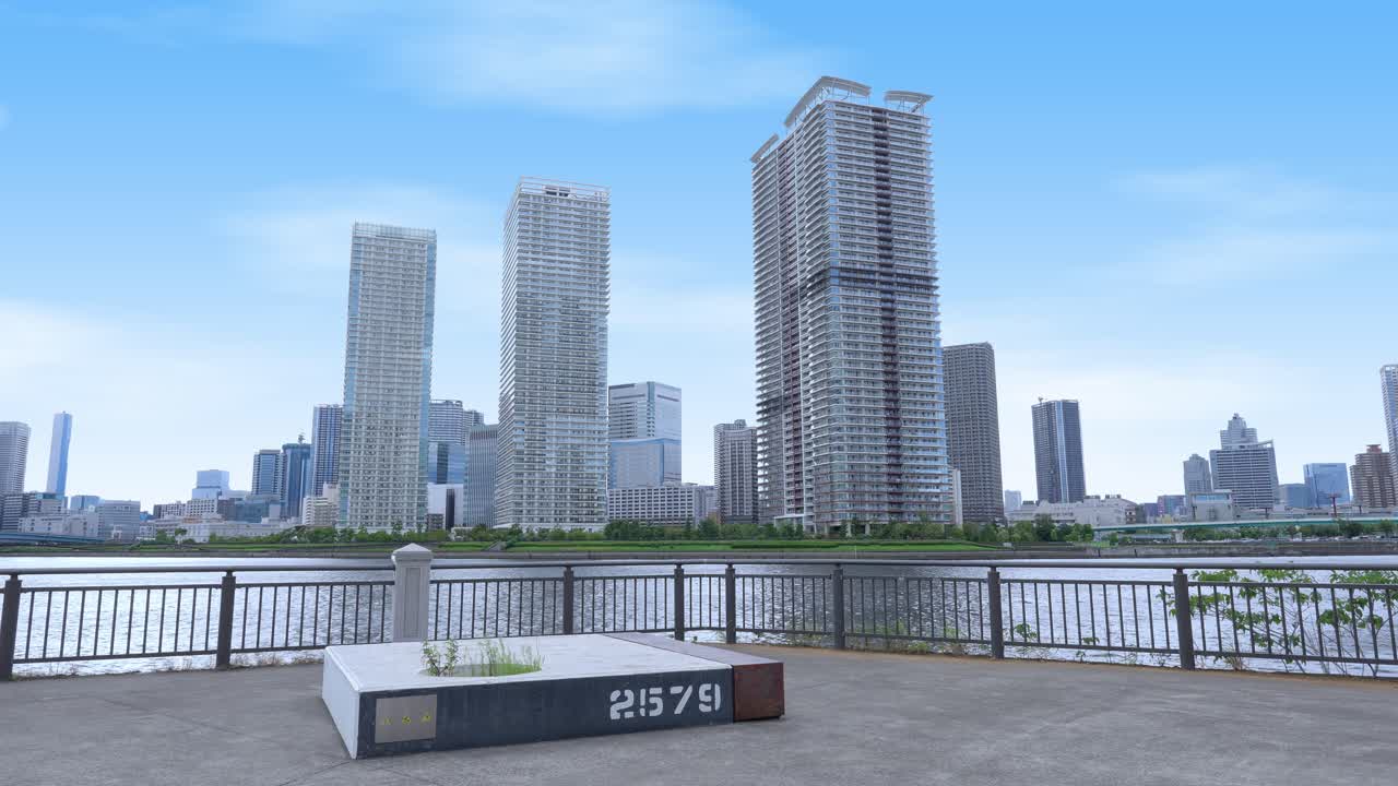A peaceful wide shot of the Tokyo skyline from a waterfront promenade with modern high-rise buildings and a clear sky