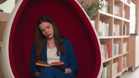 young woman reading a book in a red egg chair in a library