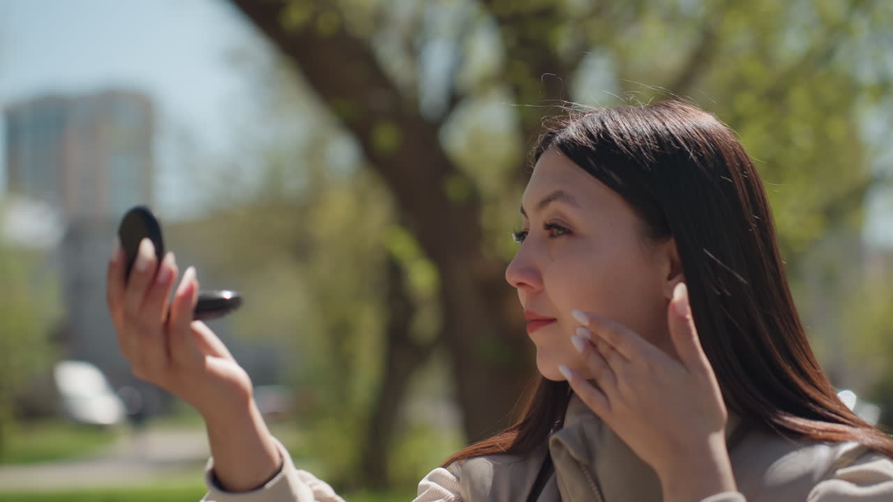 화창한 공원에서 즐기는 자기 관리, 평화로운 공원 환경에서 활기찬 나뭇잎을 배경으로 콤팩트 거울을 사용하여 외모를 가꾸며 거울로 머리를 만지는 편안한 여성