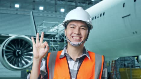 primer plano de un ingeniero masculino asiático con casco de seguridad de pie con el avión en el hangar. sonriendo a la cámara y mostrando el gesto de "ok" a la cámara mientras el mantenimiento del avión