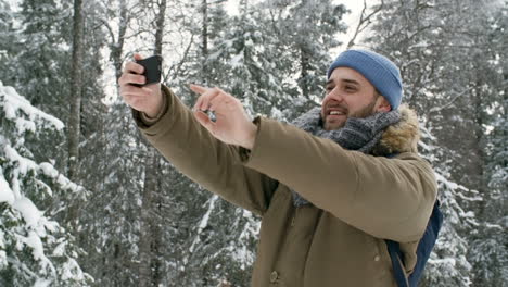 Hombre-Guapo-Alegre-Videollamada-En-El-Teléfono-Inteligente,-Saludando-A-La-Cámara,-Sonriendo-Y-Contando-Sobre-La-Hermosa-Naturaleza-Invernal-En-El-Parque-Nacional-Mientras-Camina-En-Un-Día-Nevado