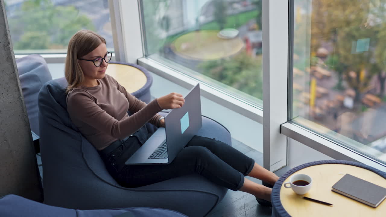 Thoughtful young lady sitting near big window opens her laptop and starts work. Woman typing on computer looking into window. High angle view.