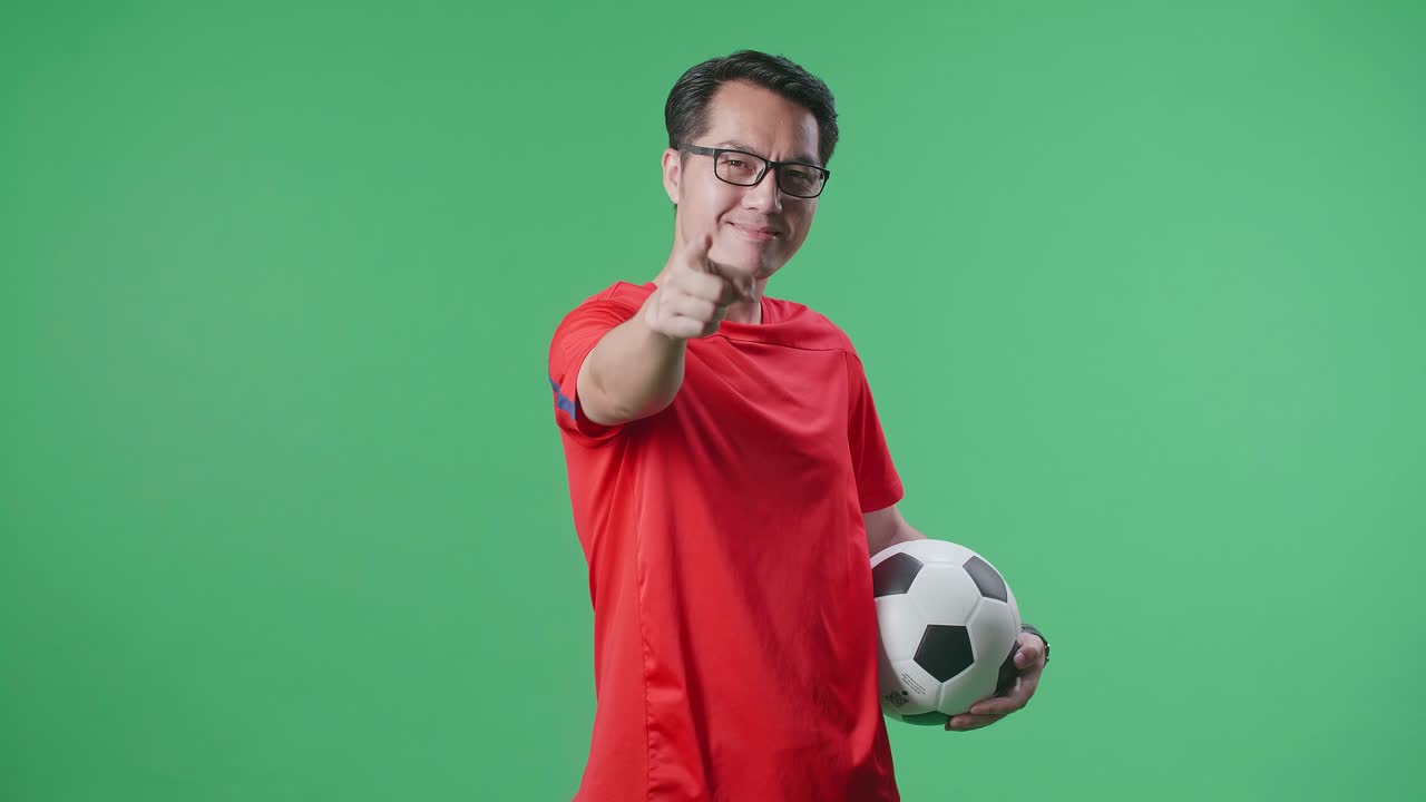 hombre asiático con una pelota poniendo la mano en el pecho y señalando hacia adelante mientras anima el fútbol en el fondo de la pantalla verde