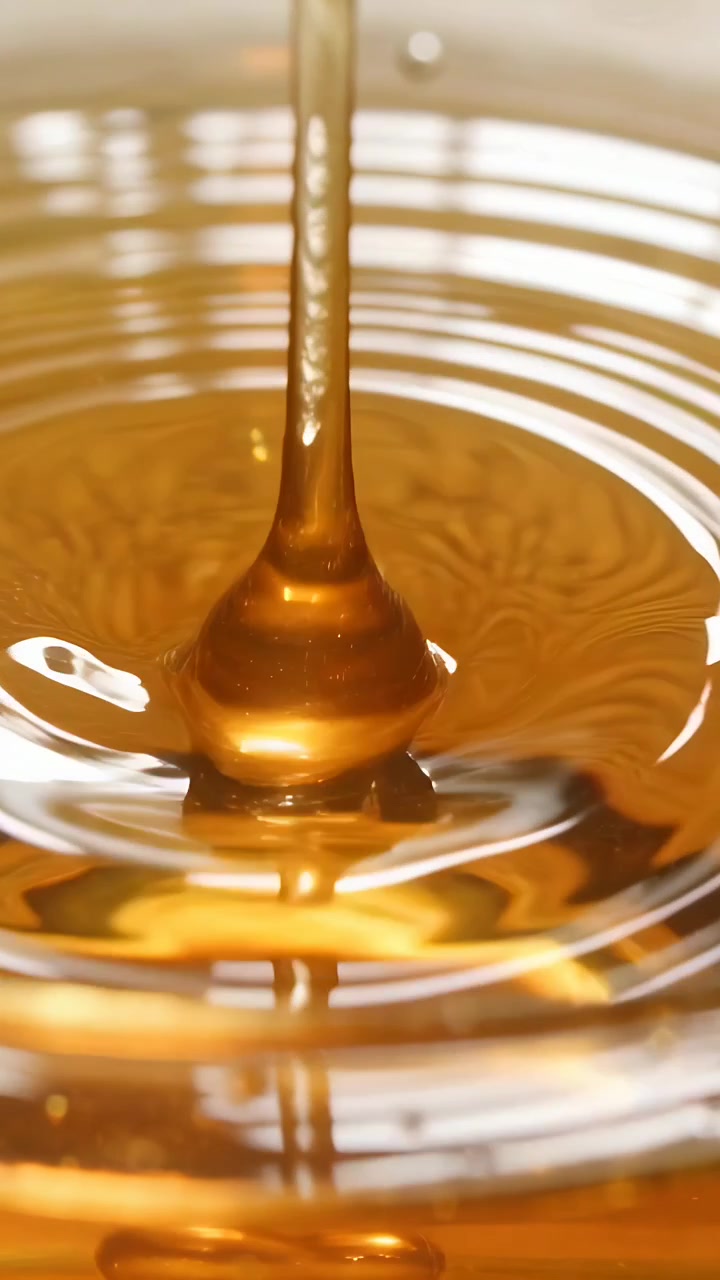 Video verticale: Goccia ambrata che si stacca e colpisce una pozza viscosa in studio, formando una colonna, una corona e delle increspature.