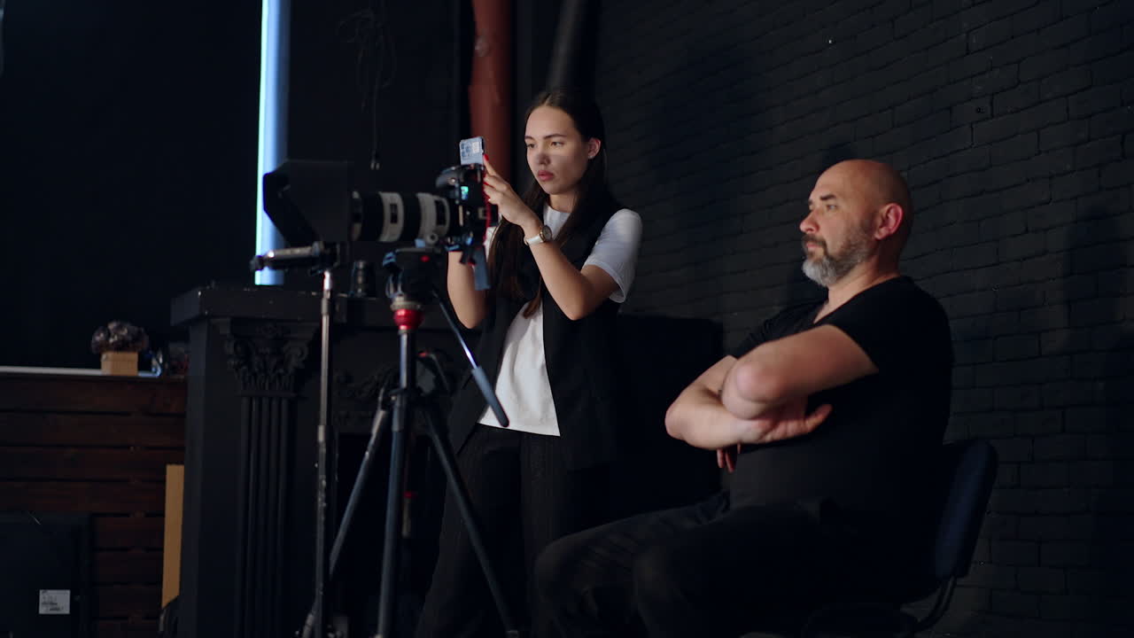 Cameraman wearing black clothes sits down behind the camera. Brunette woman in white t-shirt takes video on her phone. Studio concept