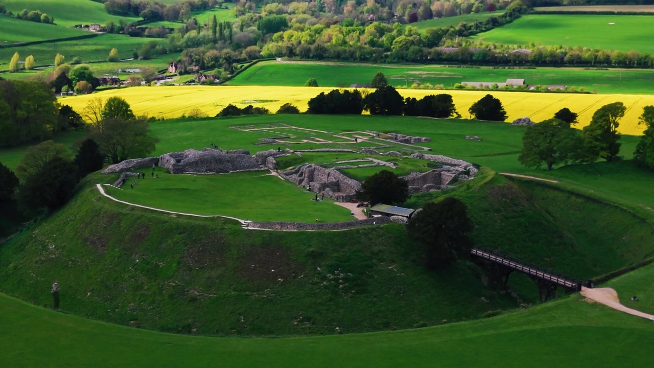 Luftaufnahme mit Drohne von Old Sarum bei Salisbury, Wiltshire, die die historischen Burgruinen und alten Wallanlagen einer Hügelfestung zeigt, umgeben von grünen Feldern, Ackerland, Bäumen und ländlicher englischer Landschaft.