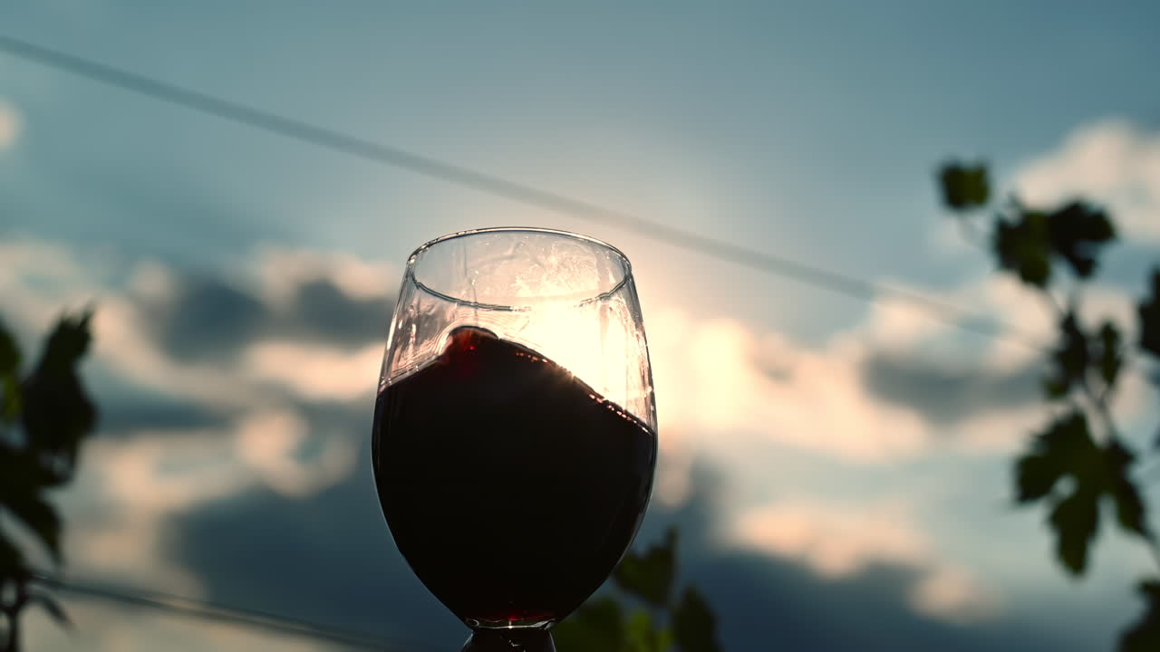 A hand holds a glass of red wine against a sunset sky in a vineyard. The sun reflects in the wine as it is poured, creating a magical atmosphere full of color and warmth