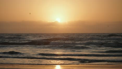 Sonne-Späht-Hinter-Wolken,-Während-Der-Vogel-Durch-Die-Goldene-Stunde-Fliegt-Sonnenaufgang-Oder-Sonnenuntergang-Am-Strand-Von-Texas-Mit-Wellen,-Die-An-Die-Küste-Krachen