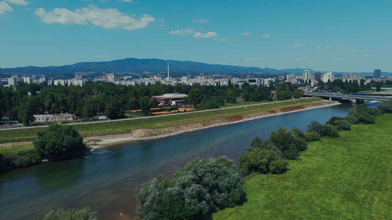 aerial - calm river flows past green banks and urban skyline of Zagreb Croatia on sunny day
