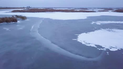 Aerial-view-of-frozen-lake-Liepaja-during-the-winter,-blue-ice-with-cracks,-dry-yellowed-reed-islands,-overcast-winter-day,-wide-drone-shot-moving-forward-low-over-the-surface