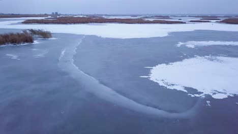 aerial view of frozen lake liepaja during the winter, blue ice with cracks, dry yellowed reed islands, overcast winter day, wide drone shot moving forward low over the surface