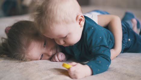 girl swings legs hugging brother with yellow toy on bed