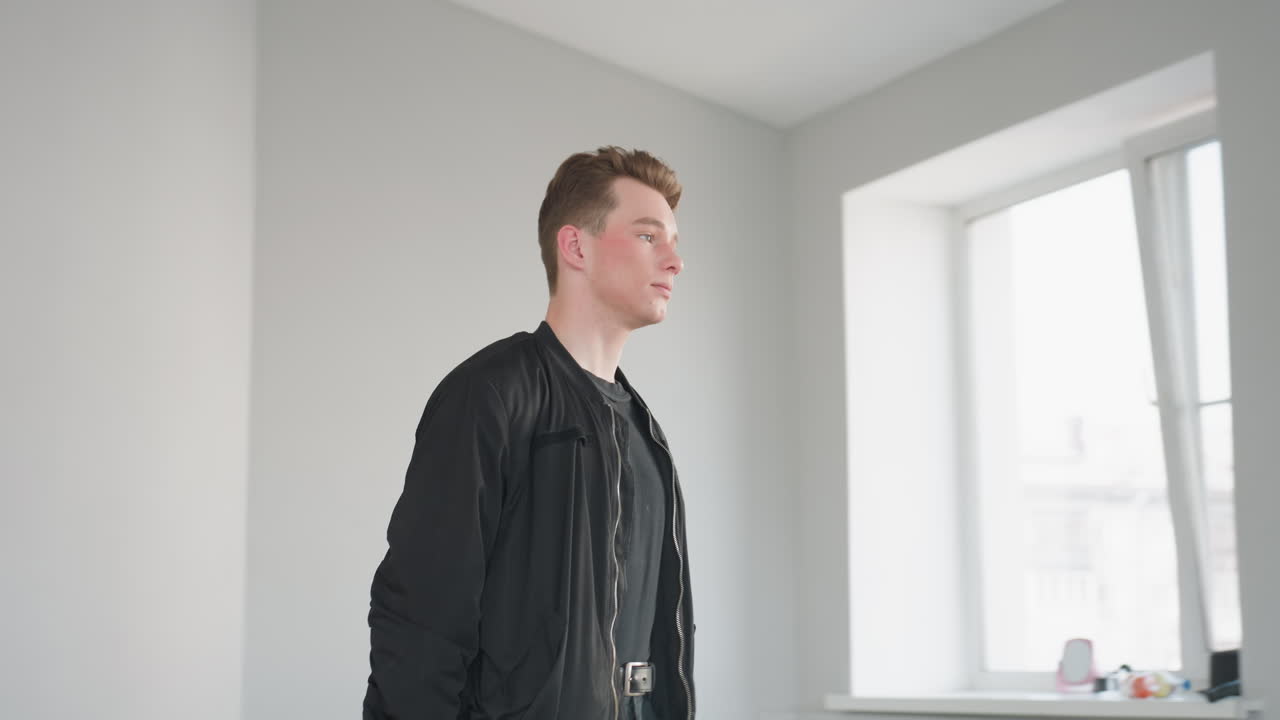 Teenager wearing black jacket and belt walks forward inside bright room pulling wheeled travel bag by handle to front, soft daylight through large windows illuminates neutral minimalist interior