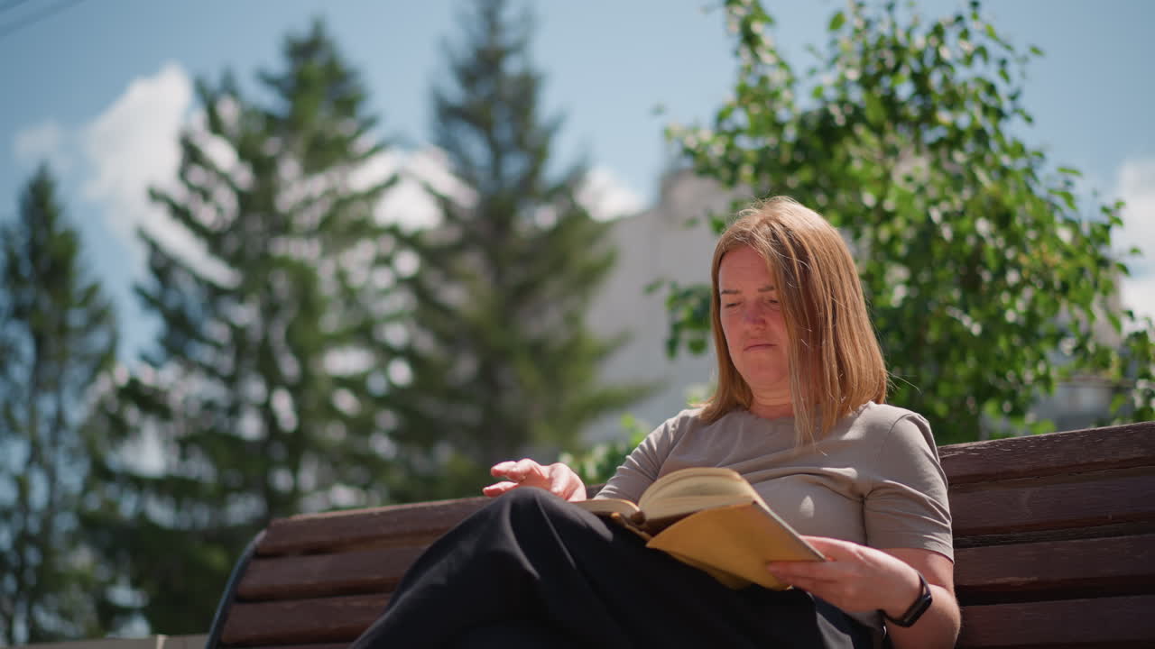 Frau sitzt auf einer Holzbank und liest ein Buch unter hellem Sommerlicht, umgeben von grünen Bäumen und klarem Himmel, ruhige und friedliche Outdoor-Atmosphäre, die einen Moment stiller Besinnung festhält
